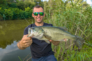 Man holding a large fish by a body of water with greenery in the background